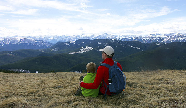 A young child (left) and adult (right) sit on a grassy patch overlooking a mountain valley. Their backs are facing the camera.