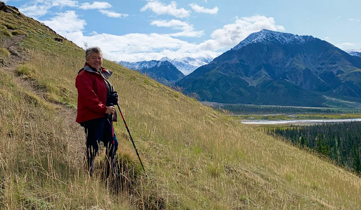 A person standing on a grassy slope smiling at the camera. In the background is a mountain valley in the Yukon, Canada.