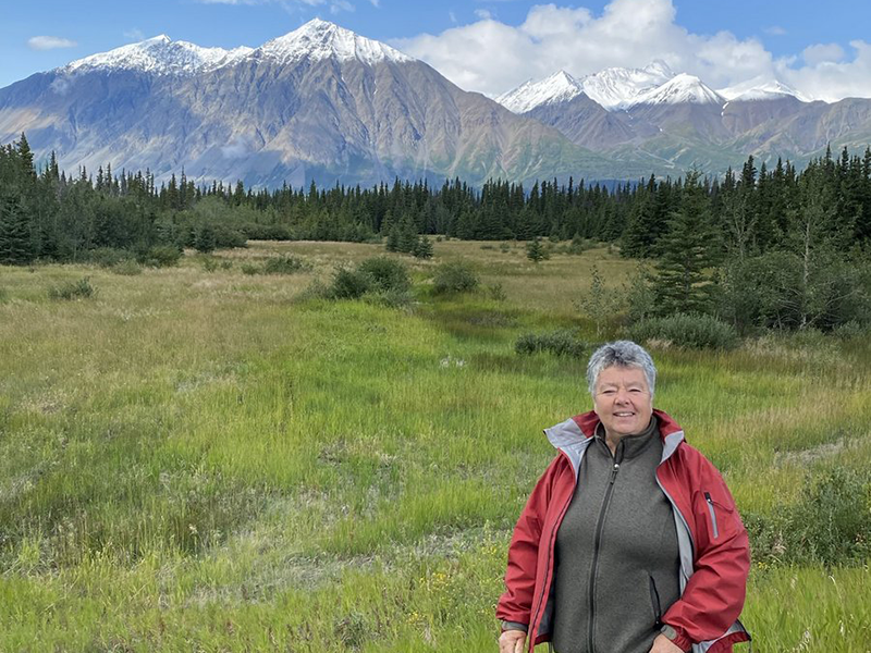Y2Y donor Maureen stands smiling at the camera with a mountain range, forest, and green grassy field in the background. This photo was taken in the Yukon, Canada.
