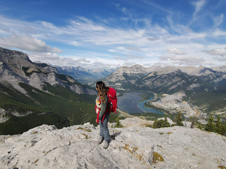 A woman stands on a cliff overlooking a large mountain and lake landscape