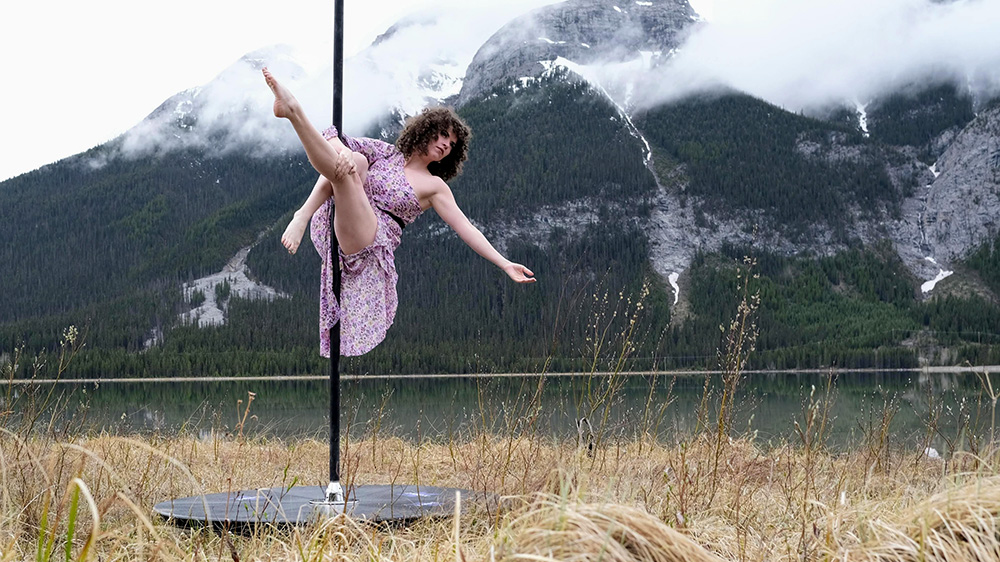 A woman with a pink chiffon dress dances on a free-standing pole in the Canadian Rockies. The background is a cloudy mountain lake and forest scene