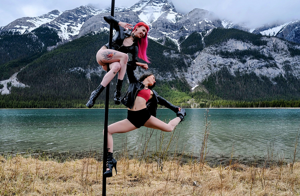Two women artistically hang as they dance on a free-standing pole in the Canadian Rockies. The background is a cloudy mountain lake and forest scene