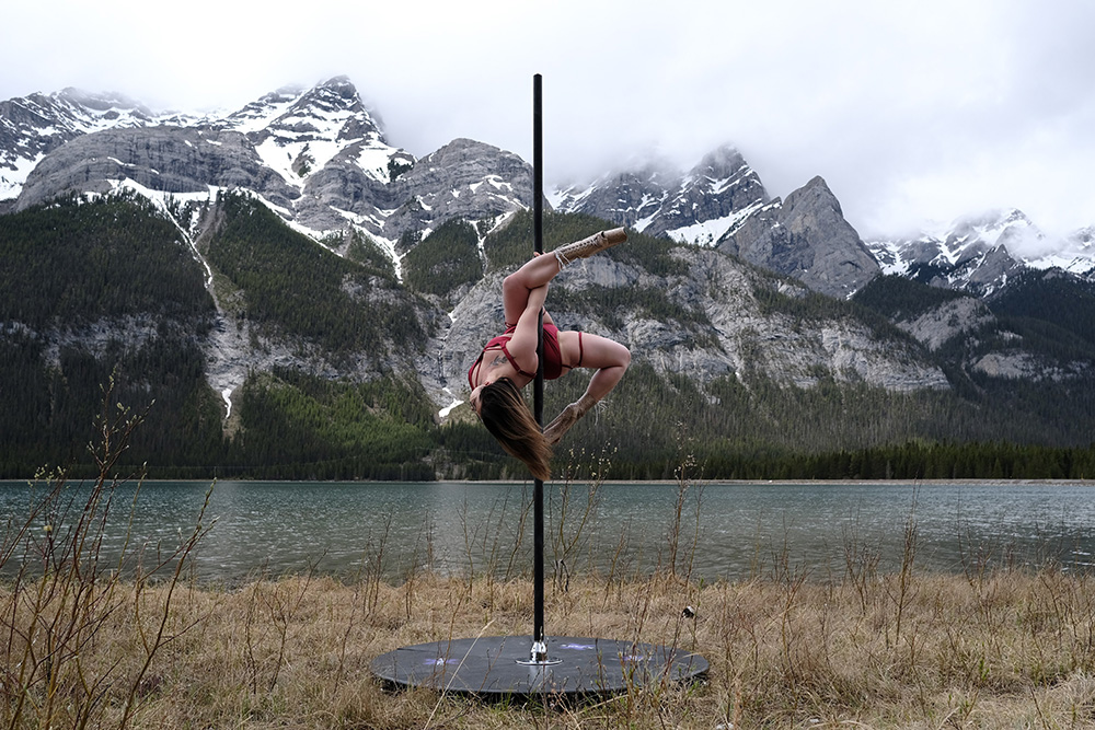 A woman slides upside down as she dances on a free-standing pole in the Canadian Rockies. The background is a cloudy mountain lake and forest scene