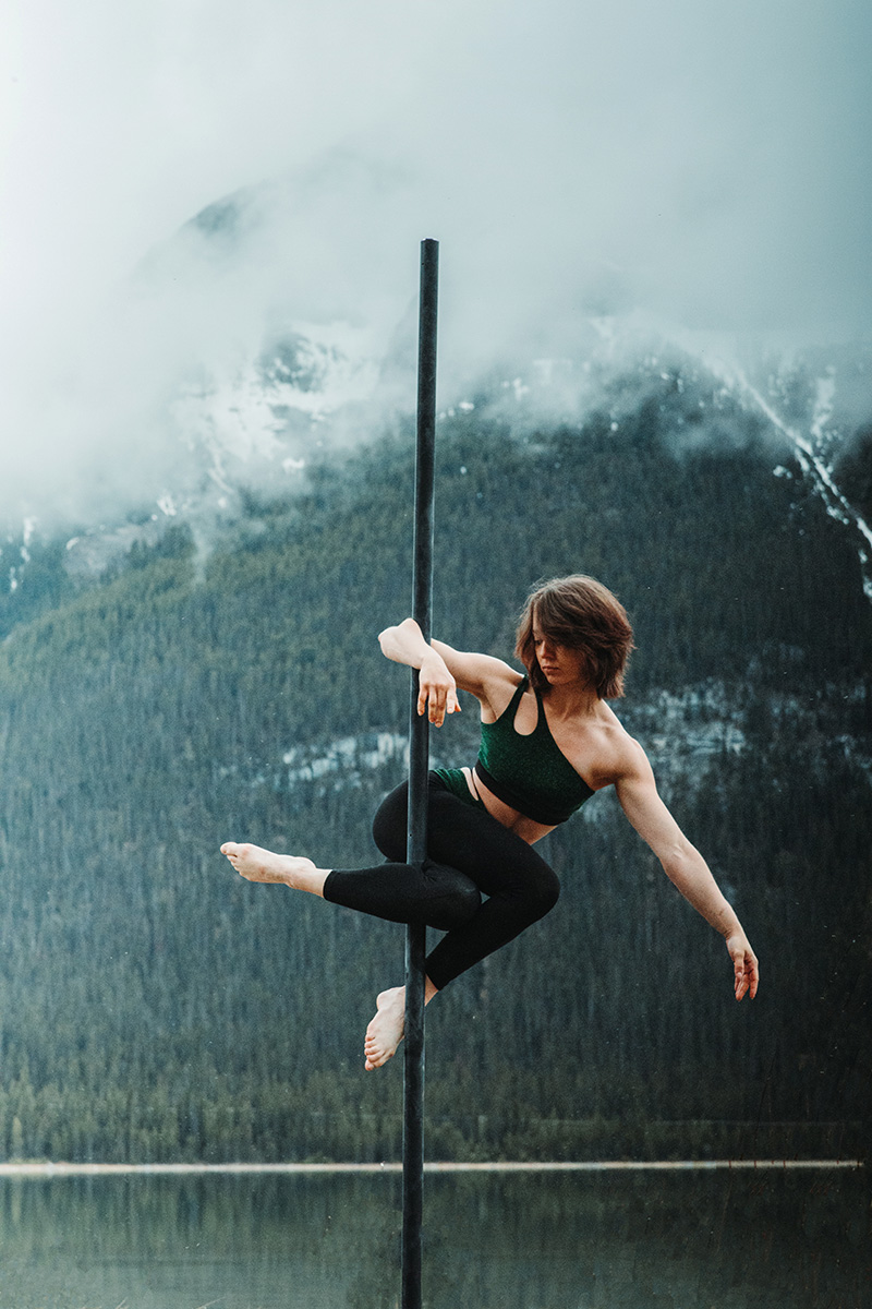 A woman dances on a free-standing pole in the Canadian Rockies. The background is a cloudy mountain lake and forest scene