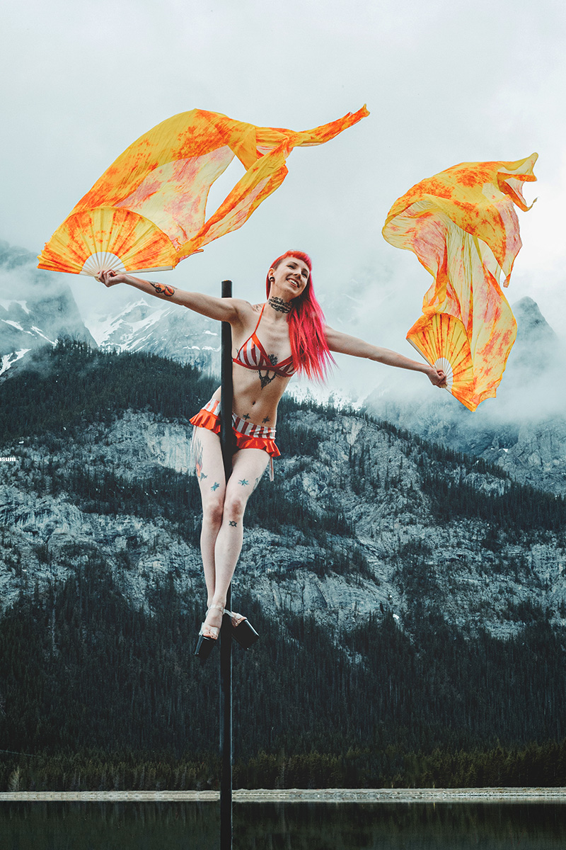 A woman dances on a free-standing pole in the Canadian Rockies. She is wearing a red outfit with decorative flags that look like flames.