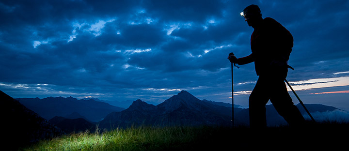 A person wearing a headlamp and using hiking poles hikes at dusk in the mountains
