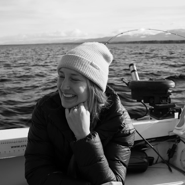 Black-and-white photo of Tenaya Lynx sitting on a boat, smiling and has her chin rested on her fist. She is wearing a light-colored toque. 