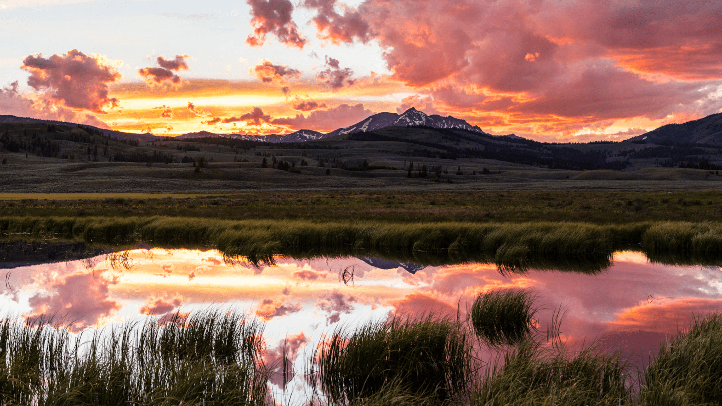 Behind a mountain in the distance is a vibrant pink, yellow and orange sunset on a cloudy evening, reflecting off a small body of water in the foreground.