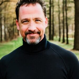 A man with short cropped dark hair and a goatee in a black turtleneck stands in a forest