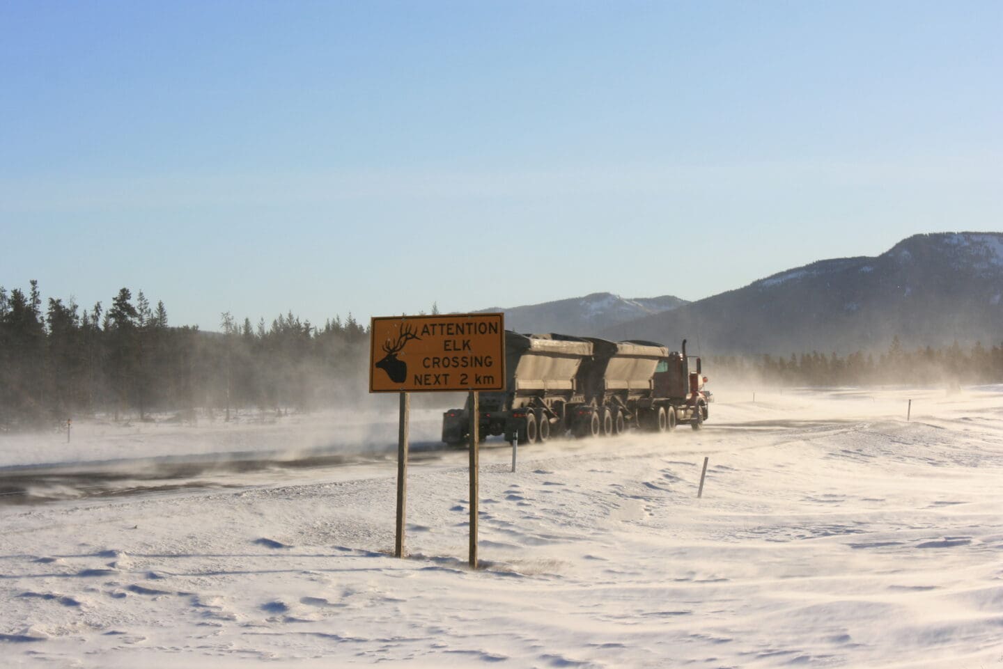 An image of a truck travelling on a windy snowy highway with a sign saying: Attention elk crossing next 2 km