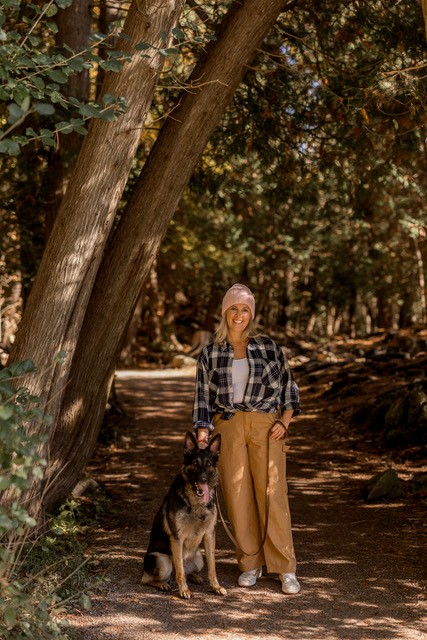 A women with blond hair smiles at the camera, walking down a path in a shaded forest. She is wearing a light pink hat and a black and white plaid shirt.