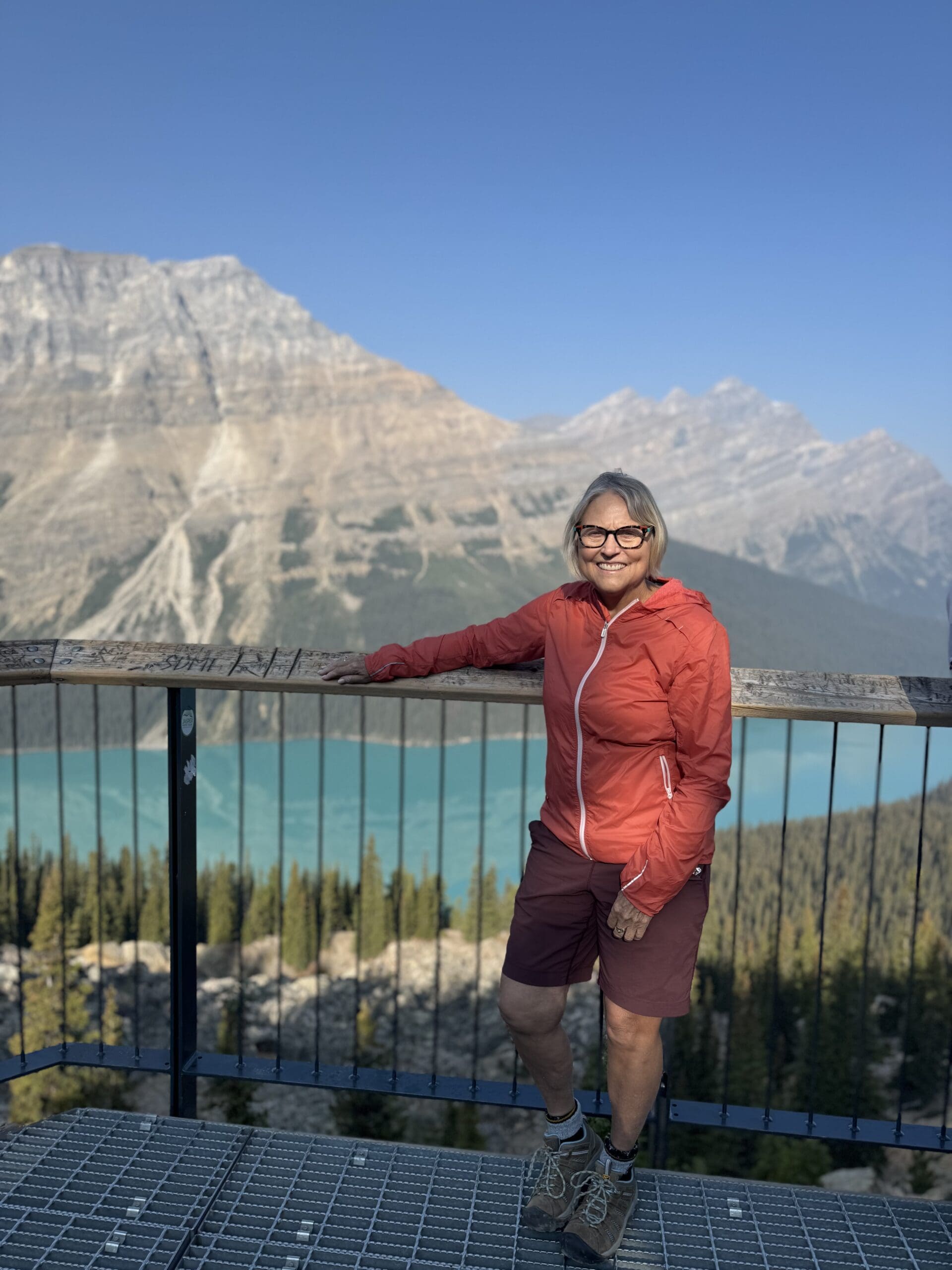 A light-haired women smiles at the camera, with mountains and a blue-ish green alpine lack in the background. The women is wearing glasses and and orange windbreaker jacket.