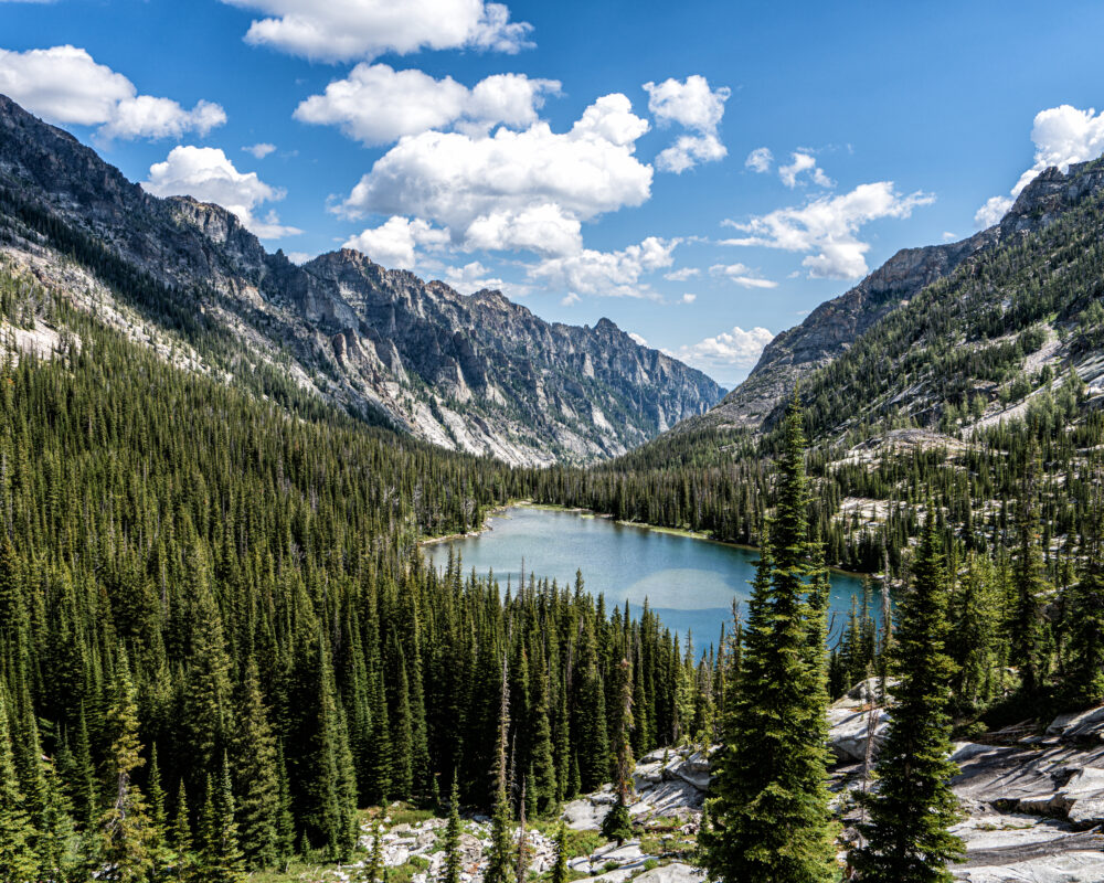 Rugged mountains surround a small alpine lake. The slopes closer to the lake are covered in a forest of coniferous trees.