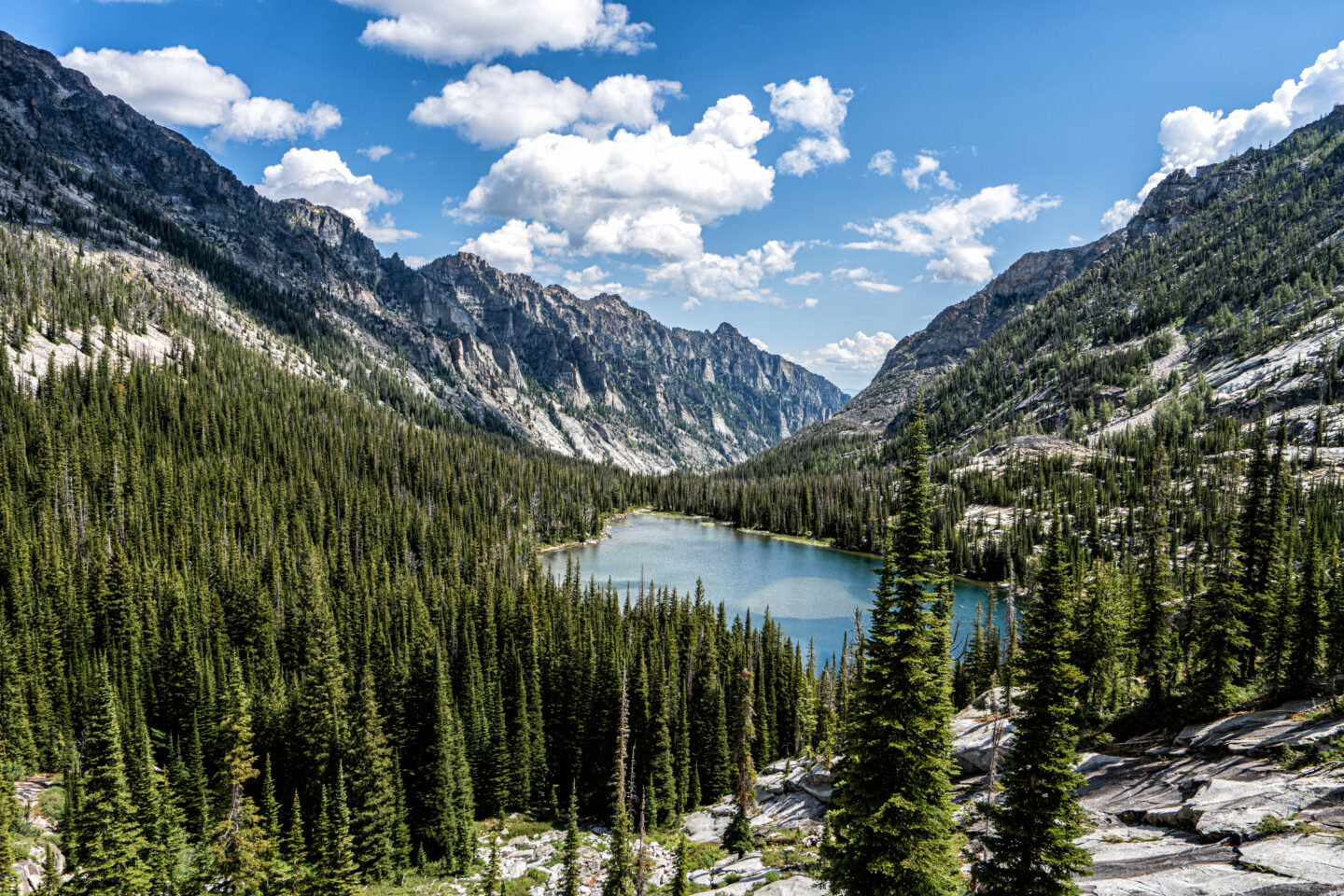 Rugged mountains surround a small alpine lake. The slopes closer to the lake are covered in a forest of coniferous trees.