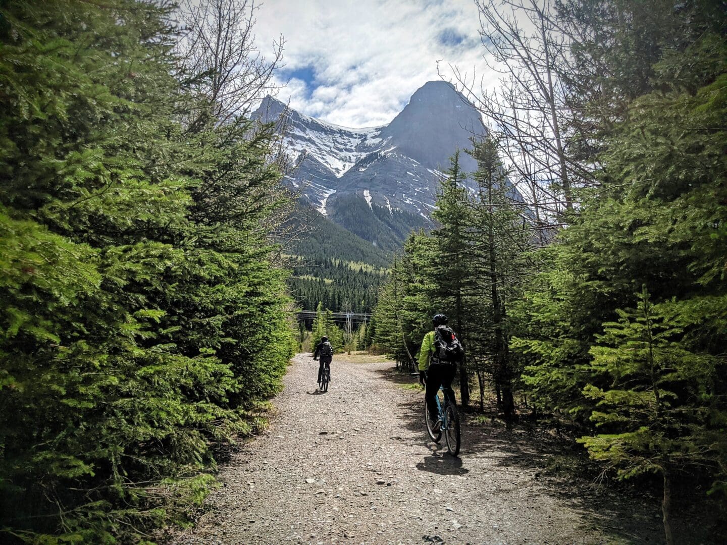 People ride mountain bikes down a dirt path towards Mount Lawrence Grassi (Ehagay Nakoda) in Alberta's Bow Valley