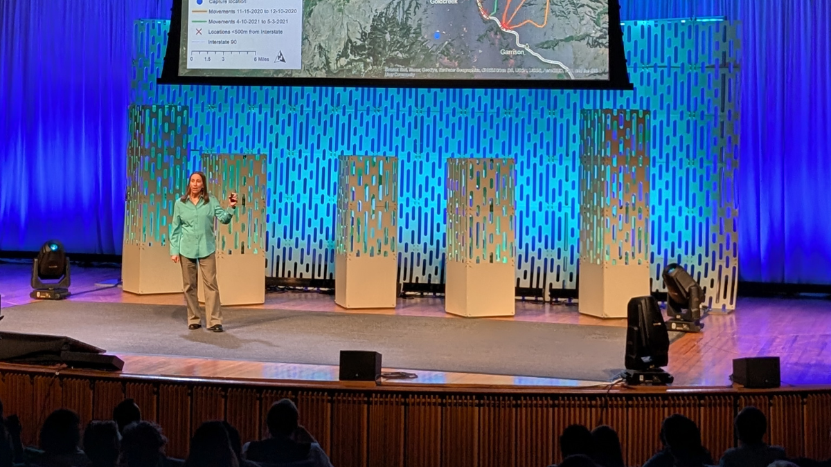 Dr. Jodi Hilty stands onstage at the TEDxBoston talk, with a map illustrating attempted wildlife crossings of a highway behind her. She is talking to the crowd.